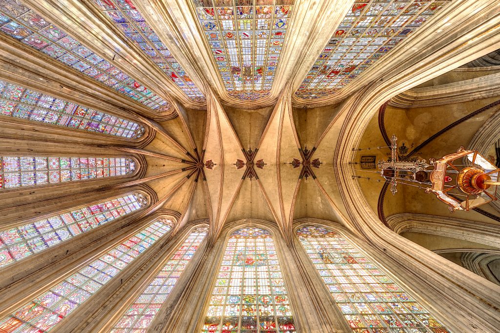 View of the vaulted ceiling of Notre-Dame church in Le Sablon, with coloured stained glass windows and a large suspended chandelier.